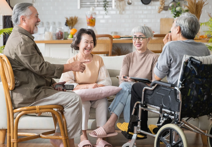 Four older adults laughing and socializing at home, including a person using a wheelchair.