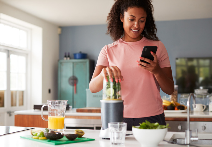 Woman using her smartphone while preparing a green smoothie with fresh ingredients in her kitchen.